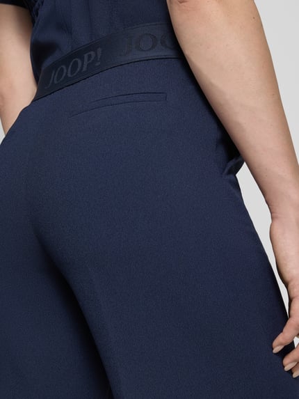 Frauenmodel trägt dunkelblaue Straight Fit Stoffhose mit Logo-Detail von JOOP!, elastischer Bund, feine Textur, Nahaufnahme Hüfte. Modell 'Paulien', Damen Stoffhose, Ziernaht, Bundweite 33 cm, Innenbeinlänge 81 cm.