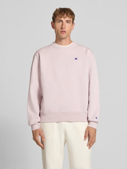 Männermodel trägt Sweatshirt mit Label-Details von Champion in blassem Rosa aus Baumwoll-Mix, lockere Passform, Rundhalsausschnitt, überschnittene Schultern, Ganzkörperansicht von vorne.