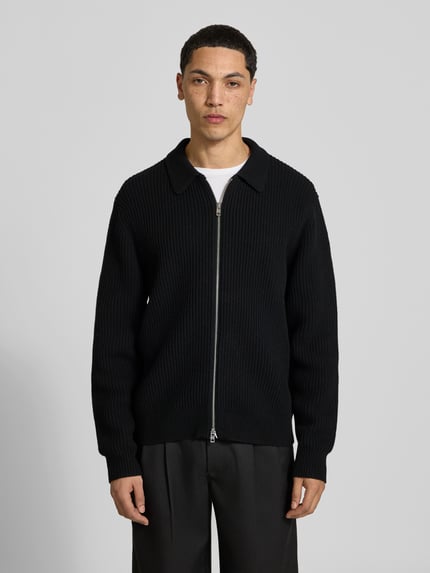 Männermodel trägt Cardigan aus reiner Wolle von Filippa K in Schwarz, Ripp-Optik, Zweiwege-Reißverschluss, lockere Passform, Ganzkörperansicht von vorne.