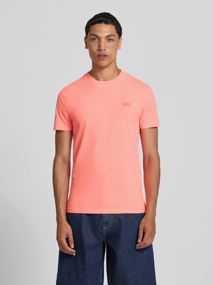 Männermodel trägt T-Shirt mit Rundhalsausschnitt und Label-Stitching von Superdry in Koralle, Reine Baumwolle, Slim-Fit, Ganzkörperansicht von vorne. Rückenlänge bei Größe M: 66 cm.