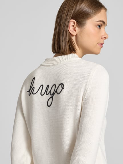 Vrouwenmodel draagt Regular Fit Pullover aus reiner Baumwolle Modell 'Saje_B' van HUGO Blue in gebroken wit met geborduurde zwarte tekst, detailopname schouder en rug.