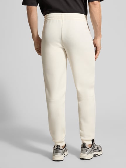 Männermodel trägt Sweatpants mit Viskose-Anteil Modell 'TRAIN PREMIUM SHIELD' von EA7 Emporio Armani in Creme, Ganzkörperansicht Rückansicht.