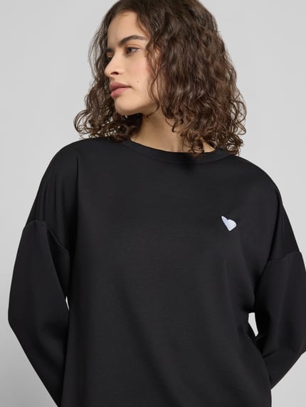 Kobiece modelka nosi czarny sweter z wyhaftowanym sercem Relaxed Fit Pullover mit Herz-Stickerei Modell 'TALIA' od Pieces, efekt: jednolity kolor, krój relaxed fit, okrągły dekolt, długi rękaw, wyhaftowane serce, zbliżenie na górną część ciała.