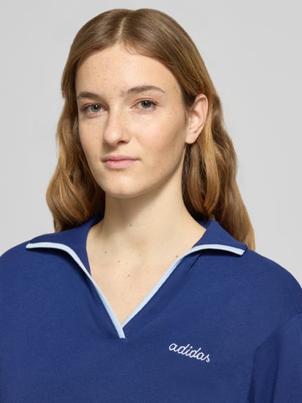 Frauenmodel trägt Poloshirt mit Label-Stitching von adidas Sportswear in Dunkelblau, reine Baumwolle, gerippte Ärmelabschlüsse, Nahaufnahme Oberkörper.