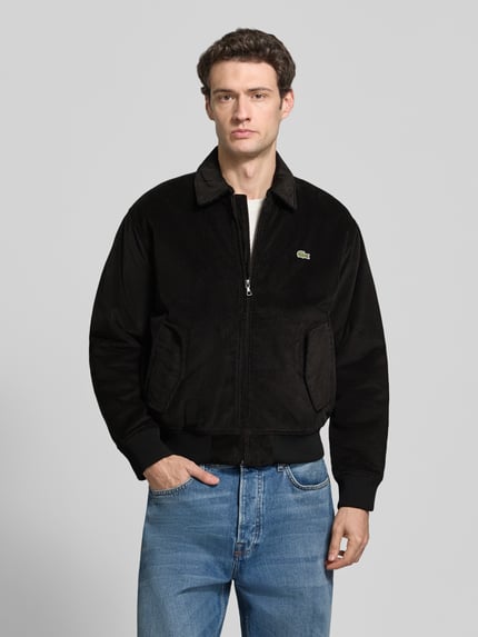 Männermodel trägt Bomberjacke mit Cordstruktur von LACOSTE in Schwarz mit Logo-Patch und seitlichen Taschen, Ganzkörperansicht von vorne.