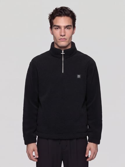 Männermodel trägt Sweatshirt mit Logo-Patch Modell 'Derylong' von HUGO in Schwarz, Fleece-Struktur, hochgeschlossener Troyerkragen, Ganzkörperansicht von vorne.