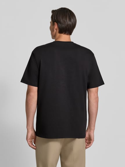 Męski model trägt Regular Fit T-Shirt aus reiner Baumwolle Modell 'Oliver' von Wood Wood, czarny kolor, gładka struktura, damski t-shirt, krój regular fit, wyhaftowane logo, Długość tyłu w rozmiarze M: 73 cm, Ganzkörperansicht Rückansicht.