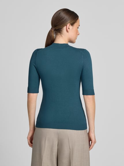 Frauenmodel trägt Slim Fit Strickpullover aus Viskose-Mix von COMMA in Petrol mit gerippten Abschlüssen, Halbarm, Ganzkörperansicht Rückansicht.
