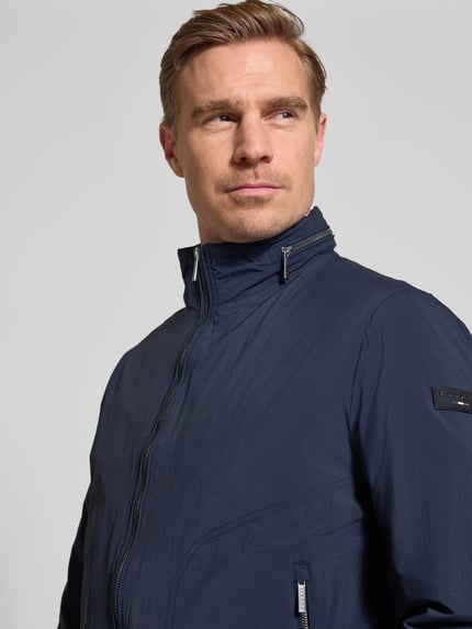 Männermodel trägt Regular Fit Jacke mit Stehkragen von bugatti in Dunkelblau mit Zweiwege-Reißverschluss, Logo-Detail, Nahaufnahme Oberkörper.