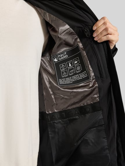 Frauenmodel trägt Jacke mit abnehmbarer Kapuze Modell 'ECLIPSE' von NAUTGÅRD in Schwarz, glänzendes Innenfutter mit Label, Nahaufnahme Oberkörper. Kontext: Damen Jacke, gerader Schnitt, Zweiwege-Reißverschluss, Seitentaschen mit Druckknopf, gestrickte Ärmelabschlüsse, Rückenlänge 65 cm, Schulterbreite 41 cm, Brustweite 51 cm, Ärmellänge 64 cm.