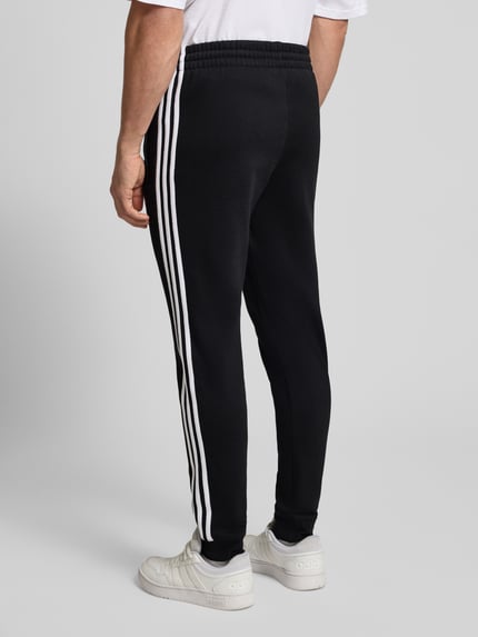 Männermodel draagt Sweatpants mit elastischem Bund van adidas Originals, zwart met witte strepen, elastische tailleband, losse pasvorm, Ganzkörperansicht Rückansicht.