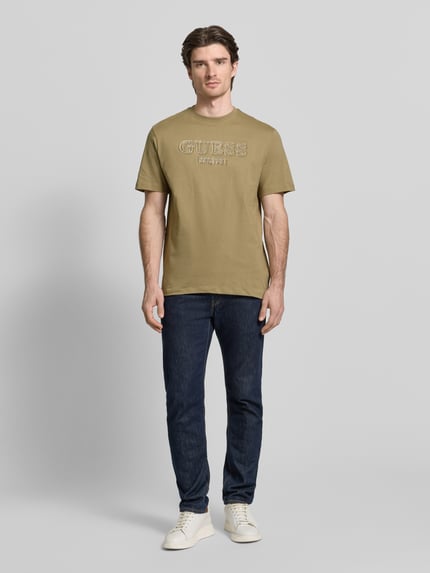 Männermodel trägt T-Shirt mit Label-Detail von Guess aus reiner Baumwolle in Oliv, Regular Fit, gerippter Rundhalsausschnitt, Ganzkörperansicht von vorne.