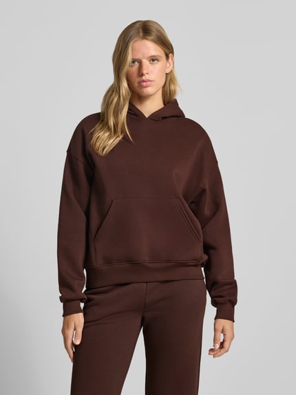 Frauenmodel trägt dunkelbraunen Relaxed Fit Hoodie aus Baumwoll-Mix Modell 'Iconic' von Gina Tricot, Känguru-Tasche, elastische Abschlüsse, Ganzkörperansicht von vorne.
