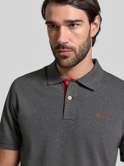 Męski model nosi Regular Fit Poloshirt mit Logo-Stitching od Gant, elastyczna bawełna, szary kolor, krótkie rękawy, haftowane logo, krój regular fit, zbliżenie na górną część ciała.