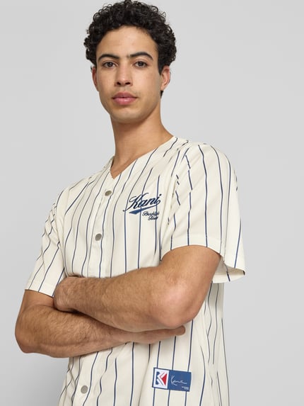 Männermodel trägt Regular Fit Freizeithemd mit Logo-Stitching von KARL KANI in Creme mit dunkelblauen Streifen, Kurzärmelig, Nahaufnahme Oberkörper. Rückenlänge 64 cm, Schulterbreite 45 cm, Ärmellänge 27 cm, V-Ausschnitt, durchgehende Knopfleiste.
