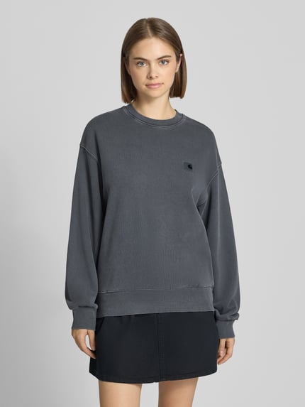 Frauenmodel trägt Relaxed Fit Sweatshirt aus reiner Baumwolle von Carhartt Work In Progress, dunkelgrau, gerippte Abschlüsse, Logo-Patch, Langarm, Ganzkörperansicht von vorne.