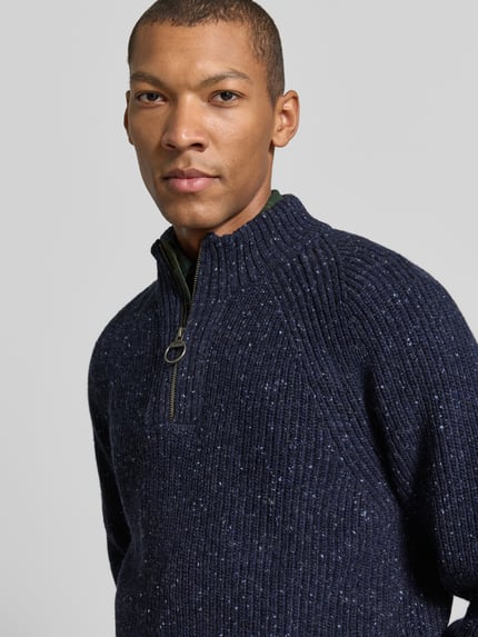 Männermodel trägt Troyer aus Woll-Mix mit Raglanärmeln von BARBOUR in dunkler Strickoptik mit Reißverschluss, Nahaufnahme Oberkörper.