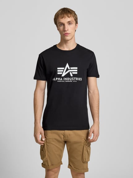 Männermodel trägt schwarzes T-Shirt mit Logo und Rundhalsausschnitt von Alpha Industries aus Baumwolle, Regular Fit, Ganzkörperansicht von vorne.