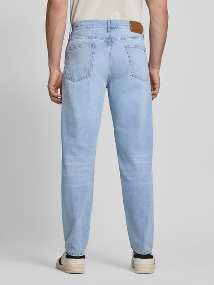 Mannenmodel draagt Tapered Fit Jeans uit puur katoen model 'HARLEM' van Tommy Hilfiger in denimlook met labelpatch, 5-pocketmodel, regular tapered pasvorm, logostitching, studs en riemlussen, knoop- en verborgen ritssluiting, lichte textuur Ganzkörperansicht Rückansicht.