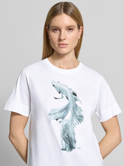 Modelka kobieca ma na sobie damski t-shirt Regular Fit T-Shirt aus reiner Baumwolle Modell 'ELPHA' von BOSS, biały z nadrukiem z motywem i logo, okrągły dekolt, krótki rękaw, dżersej z czystej bawełny, zbliżenie górnej części ciała z przodu.