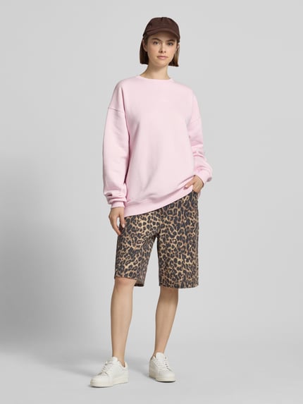 Frauenmodel trägt Oversized Sweatshirt mit Logo-Print von OH APRIL aus Baumwoll-Mix, Oversized-Fit, gerippter Rundhalsausschnitt, überschnittene Schultern, hellrosa Farbe, glatte Textur, kombiniert mit gemusterten Shorts, Ganzkörperansicht von vorne.