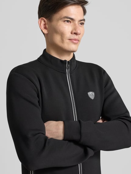 Männermodel trägt Sweatjacke mit Reißverschluss von EA7 Emporio Armani, Regular-Fit, gerippte Abschlüsse, Stehkragen, Label-Detail, Eingrifftaschen, schwarze Farbe, Nahaufnahme Oberkörper.