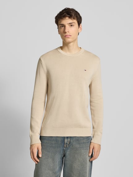 Männermodel draagt Regular Fit Strickpullover aus reiner Baumwolle van Tommy Jeans, beige effen kleur, logo detail, lange mouwen, ribboorden, Ganzkörperansicht van voren.