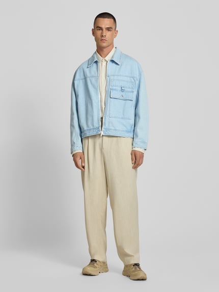 Männermodel trägt Jeansjacke aus reiner Baumwolle von MAISON KITSUNÉ in Hellblau mit Zweiwege-Reißverschluss, Gerade Passform, Brusttasche, Umlegekragen, beige Hose, neutrale Farben, Ganzkörperansicht von vorne.
