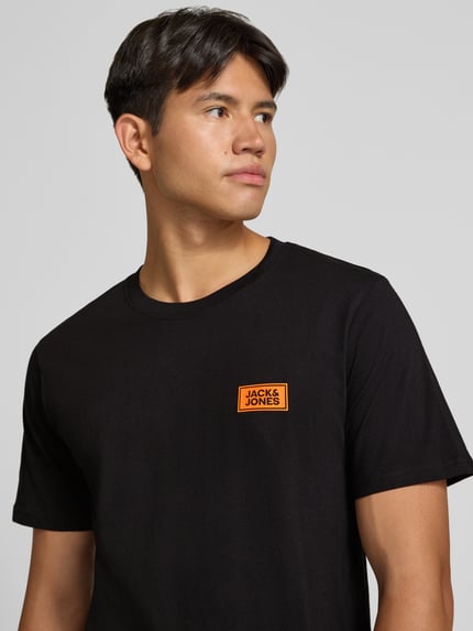 Mężczyznamodel nosi Regular Fit T-Shirt aus reiner Baumwolle mit Label-Detail von JACK & JONES, czerń, prosta faktura, dekolt okrągły, krój regular fit, bawełna, detal z logo, długość pleców w rozmiarze M: 67 cm, z przodu zbliżenie na górę ciała.