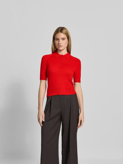 Frauenmodel trägt Poloshirt aus reiner Wolle von Victoria Beckham in Rot, Strick-Optik, Halbarm, Regular Fit, Ganzkörperansicht von vorne.