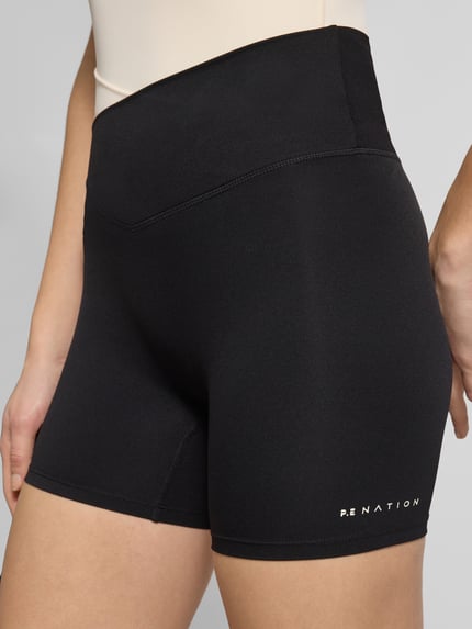 Frauenmodel trägt Slim Fit Shorts mit Logo-Print Modell 'Wander' von P.E Nation aus Elastischer-Material-Mix in Schwarz, Nahaufnahme Oberschenkel. Damen Shorts mit Elastischer-Logo-Bund, Logo-Print, Bundweite Größe S 29 cm, Innenbeinlänge 12 cm.