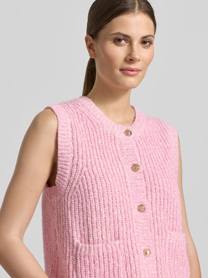 Modelka kobieca ma na sobie Strickweste Melange Effekt mit Schmuckknopf od Christian Berg Woman, różowy melanż, bawełniany mix, krój regular fit, prążkowane wykończenia, okrągły dekolt, listwa guzikowa, kieszenie, detal z guzikiem; zbliżenie na górną część ciała z przodu.