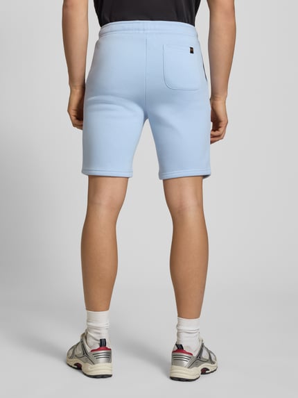 Männermodel trägt Shorts mit Eingrifftaschen und Logo-Print von Alpha Industries aus Baumwoll-Mix, Relaxed-Fit, Farbe Hellblau, Gesäßtasche, Ganzkörperansicht Rückansicht. Kontext: Herren Shorts, elastischer Bund mit Kordelzug, Bundweite 39 cm, Innenbeinlänge 24 cm Größe M.