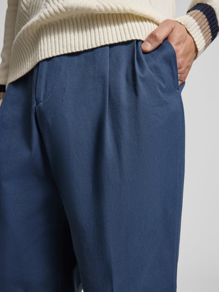 Mężczyznamodel nosi Weite Hose aus Baumwoll-Mix Modell 'PAT' od BOSS, jednokolorowe, krój relaxed fit, spodnie z szerokimi nogawkami, zakładki w pasie, wykończenie: splot twill, mieszanka bawełny, zapięcie na haftkę i ukryty zamek błyskawiczny, boczne wpuszczane kieszenie, tylne kieszenie, tekstura gładka, kolor niebieski, zbliżenie na górną część spodni.