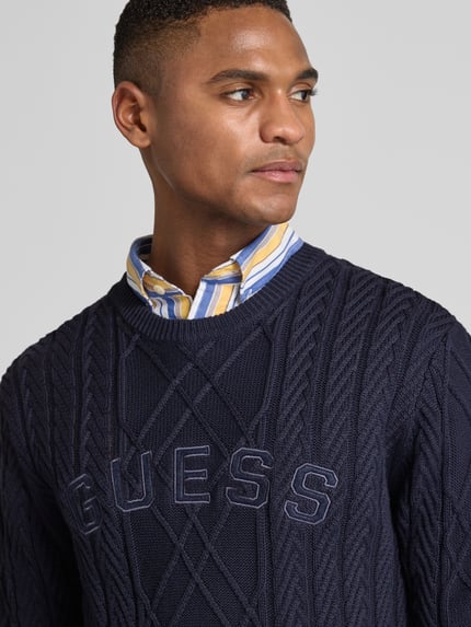 Männermodel trägt dunkelblauen Strickpullover aus Baumwoll-Modal-Mix von Guess mit Rundhalsausschnitt, Nahaufnahme Oberkörper.