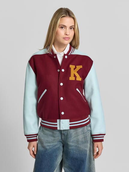 Frauenmodel trägt College-Jacke mit Logo-Patch von Kenzo, bordeaux und hellblau, Two Tone, Struktur, Kontraststreifen, Webware, Rippen, Ganzkörperansicht von vorne.