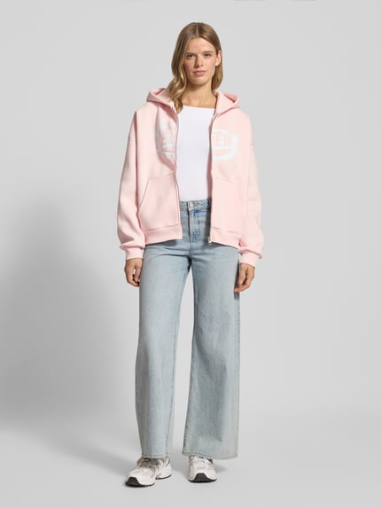 Frauenmodel trägt Sweatjacke mit Logo und Känguru-Tasche von Review in Rosa, weiche Textur, relaxed Fit, Ganzkörperansicht von vorne.