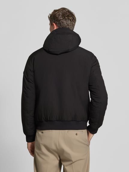 Männermodel trägt Funktionsjacke mit Kapuze Modell 'C-Caniol' von BOSS in Schwarz, strukturierte Webware, unifarben, Reißverschlusstaschen, Ganzkörperansicht Rückansicht.
