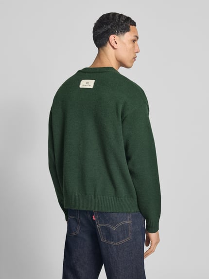 Männermodel trägt ciemnozielony Cardigan mit Logo-Stitchings und V-Ausschnitt von ANOTHER COTTON LAB, prosty krój, dekolt w serek, listwa guzikowa, wyhaftowane logo, prążkowane wykończenia, długość tyłu w rozmiarze M: 61 cm, długość rękawów w rozmiarze M: 59 cm, tkanina o gładkiej teksturze, Ganzkörperansicht Rückansicht.
