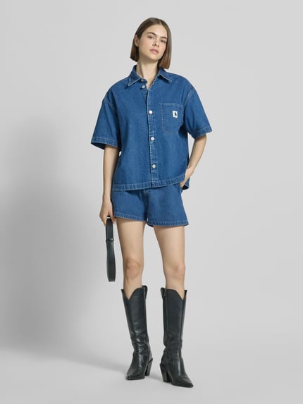 Vrouwenmodel draagt Shorts in Denim-Optik Modell 'Lova' van Carhartt Work In Progress van puur katoen, denimlook, uitlopende pasvorm, steekzakken, achterzak, knoop- en ritssluiting, bandwijdte maat S 38 cm, binnenbeenlengte 6 cm, geheel lichaamsaanzicht van voren.