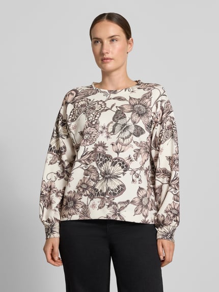 Kobiece model prezentuje Regular Fit Bluse aus Viskose-Mix von monari z mieszanki wiskozy, krój regular fit, okrągły dekolt, kwiatowy nadruk na całej powierzchni, długie rękawy, widoczne kontrasty w kolorach kremowym i brązowym, widoczne faktury materiału, widok całej sylwetki z przodu.