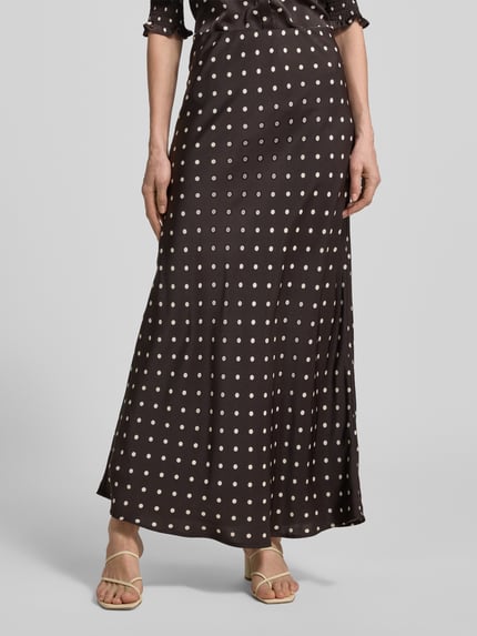 Frauenmodel trägt Maxirock aus Satin im Allover-Look Modell 'VISO DOT' von Neo Noir, braun mit weißen Punkten, A-Linie, Nahaufnahme Unterkörper. Bundweite bei Größe 36: 36 cm, Gesamtlänge bei Größe 36: 102 cm.
