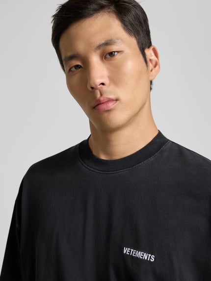 Männermodel trägt Relaxed Fit T-Shirt aus reiner Baumwolle von VETEMENTS in Schwarz mit Rundhalsausschnitt und Label-Stitching, Nahaufnahme Oberkörper.
