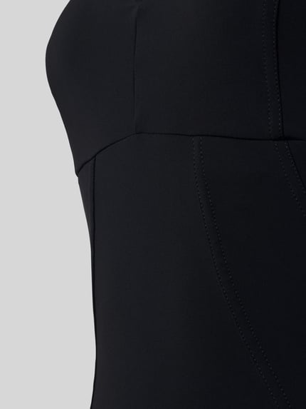 Vrouwenmodel draagt Badeanzug mit Shaping-Effekt und V-Ausschnitt Modell 'Vertigo' van Arena, zwart, gladde stof met fijne naden, shapewear, Sensitive® Fabrics, logo-print, verstelbare bandjes, chloorbestendig, uv-bescherming, sneldrogend, detailopname buikstreek.