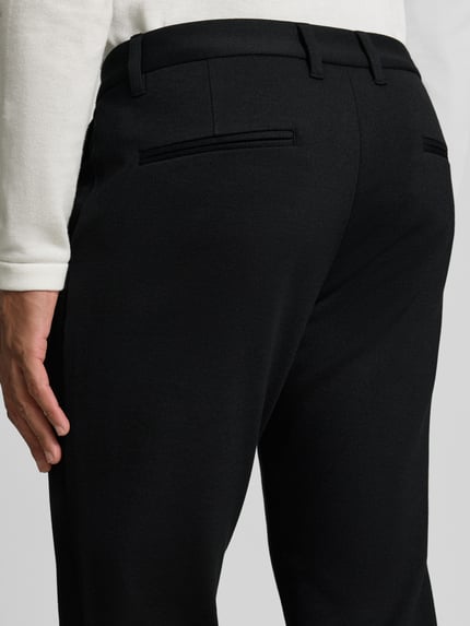 Männermodel trägt Slim Fit Stoffhose mit umgeschlagenen Hosenbeinen von Lindbergh, schwarze Farbe, Detailaufnahme Rückansicht, elastischer Material-Mix mit Viskose-Anteil, Gürtelschlaufen, Knopf- und Reißverschluss, Bundweite bei Größe M 43 cm, Innenbeinlänge 73 cm.