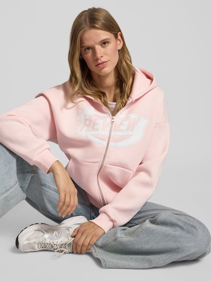 Frauenmodel trägt Sweatjacke mit Logo und Känguru-Tasche von Review in Rosa, weicher Baumwoll-Mix, Relaxed Fit, Ganzkörperansicht von vorne.