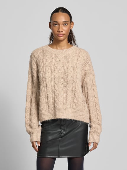 Frauenmodel trägt Oversized Fit Strickpullover mit Zopfmuster von REVIEW in Beige, grobe Struktur, kombiniert mit schwarzem Minirock, Ganzkörperansicht von vorne. Rückenlänge bei Größe S 54 cm, Ärmellänge 58 cm, elastische Abschlüsse, Rundhalsausschnitt.