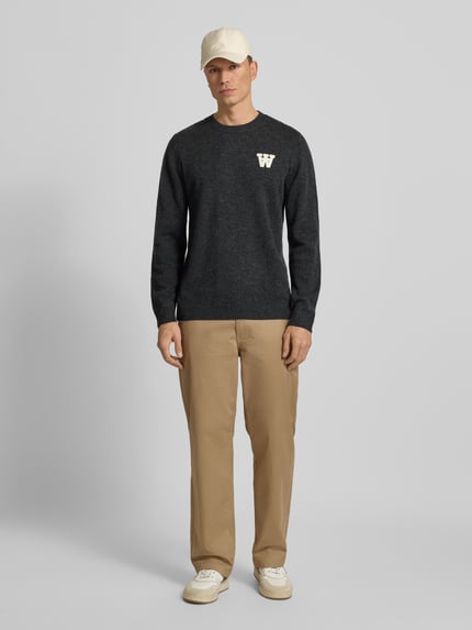 Männermodel trägt Strickpullover aus Lammwolle Modell 'Tay' von Wood Wood, dunkelgrau, Rundhalsausschnitt, Regular-Fit, Label-Stitching, Ganzkörperansicht von vorne. Kontext: Herren Strickpullover, Rückenlänge bei Größe M 72 cm, Ärmellänge 68 cm, reine Lammwolle.