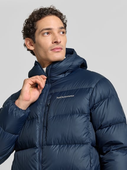 Männermodel trägt Steppjacke mit Kapuze und Wyhaftowane logo von Peak Performance, granatowy, krój regular fit, zamek błyskawiczny, Nahaufnahme górnej części ciała. Długość tyłu w rozmiarze M: 78 cm, szerokość ramion w rozmiarze M: 42 cm, szerokość w klatce piersiowej w rozmiarze M: 55 cm, długość rękawów w rozmiarze M: 68 cm.