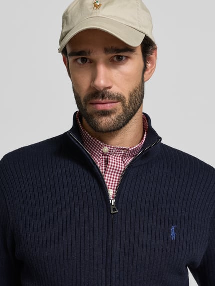 Męski model nosi Kardigan rozpinany z zamkiem błyskawicznym Polo Ralph Lauren z bawełny, krój regular fit, prążkowane wykończenia, stójka, wyhaftowane logo, wpuszczane kieszenie, kolor granatowy, długość tyłu 70 cm, długość rękawa 65 cm, zbliżenie na górną część tułowia.
